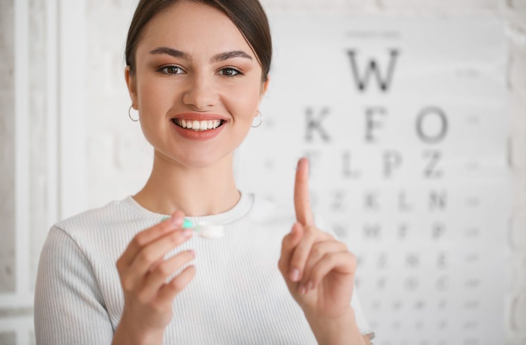 A young woman in front of Snellen chart holding a contact lens and lens case.