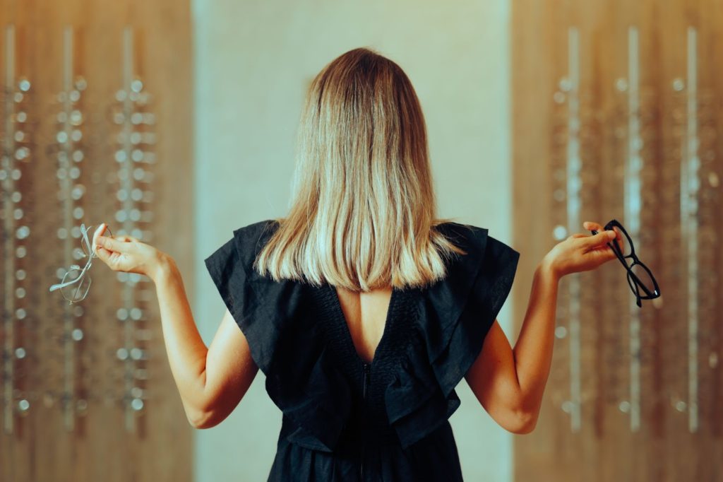 A woman with her back to the camera holds two different glasses in each hand, in front of an out-of-focus wall of glasses.