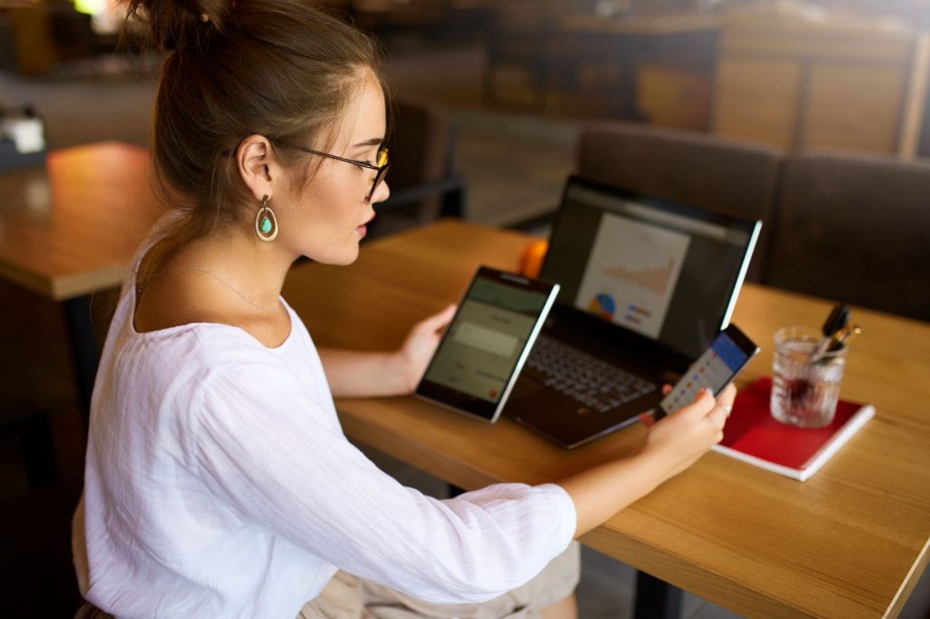 A woman in glasses holds a phone and tablet in each hand while a computer sits open on the table in front of her.