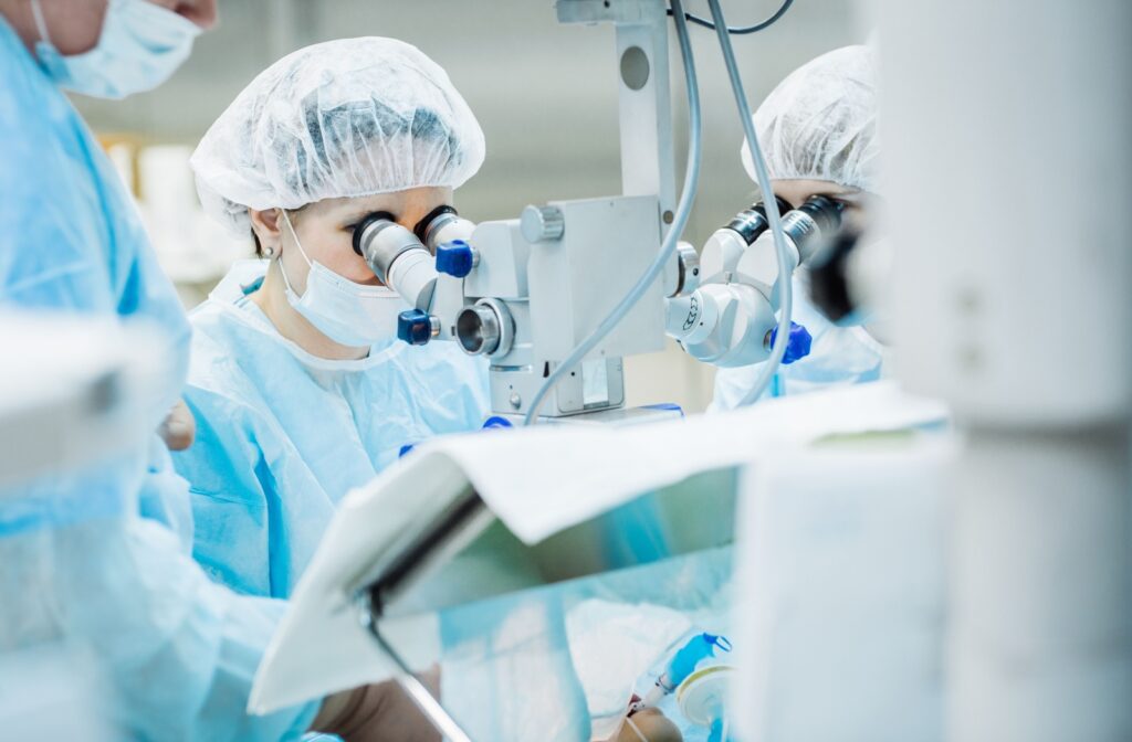 Medical professionals wearing surgical gowns, masks, and hair covers performing eye surgery using a specialized operating microscope in a sterile environment, likely during a procedure such as cataract removal.