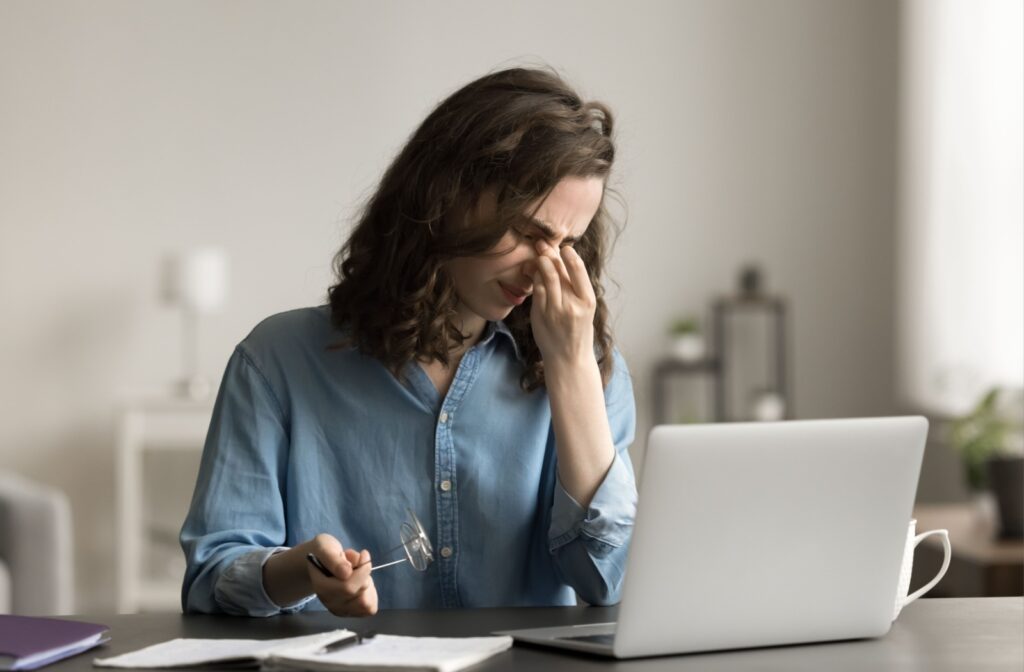 Woman sitting at a desk in front of a laptop, holding her glasses and pinching the bridge of her nose in apparent discomfort, possibly due to eye strain or a headache from prolonged screen time.