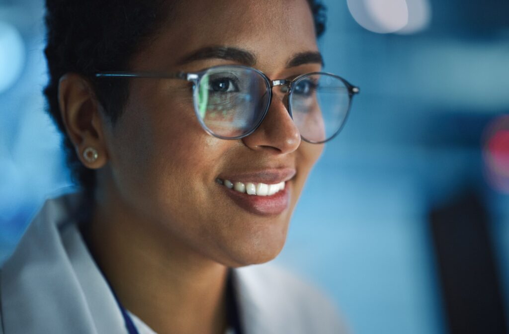 Smiling woman wearing glasses, looking at a screen with a soft blue background; reflections of digital content visible on her lenses, suggesting a professional or tech-focused environment.