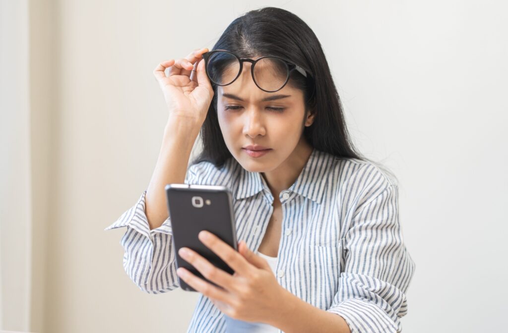 Woman experiencing blurry vision while looking at her phone, lifting her glasses and squinting in an attempt to see the screen more clearly.