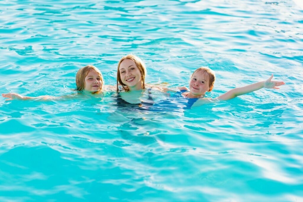 A woman and two young girls smiling and floating in a bright blue swimming pool with their arms outstretched.